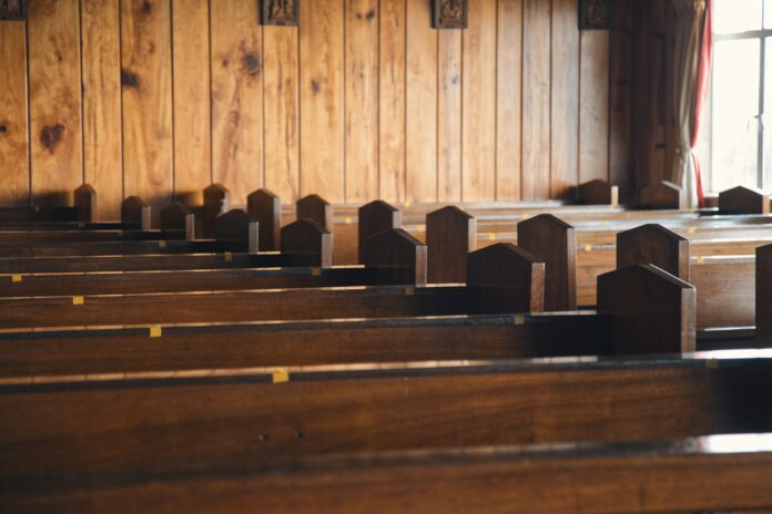 Rows of empty dark wooden church pews beside a wood-paneled wall, with a bright window at the right, an image for modesty culture in church.