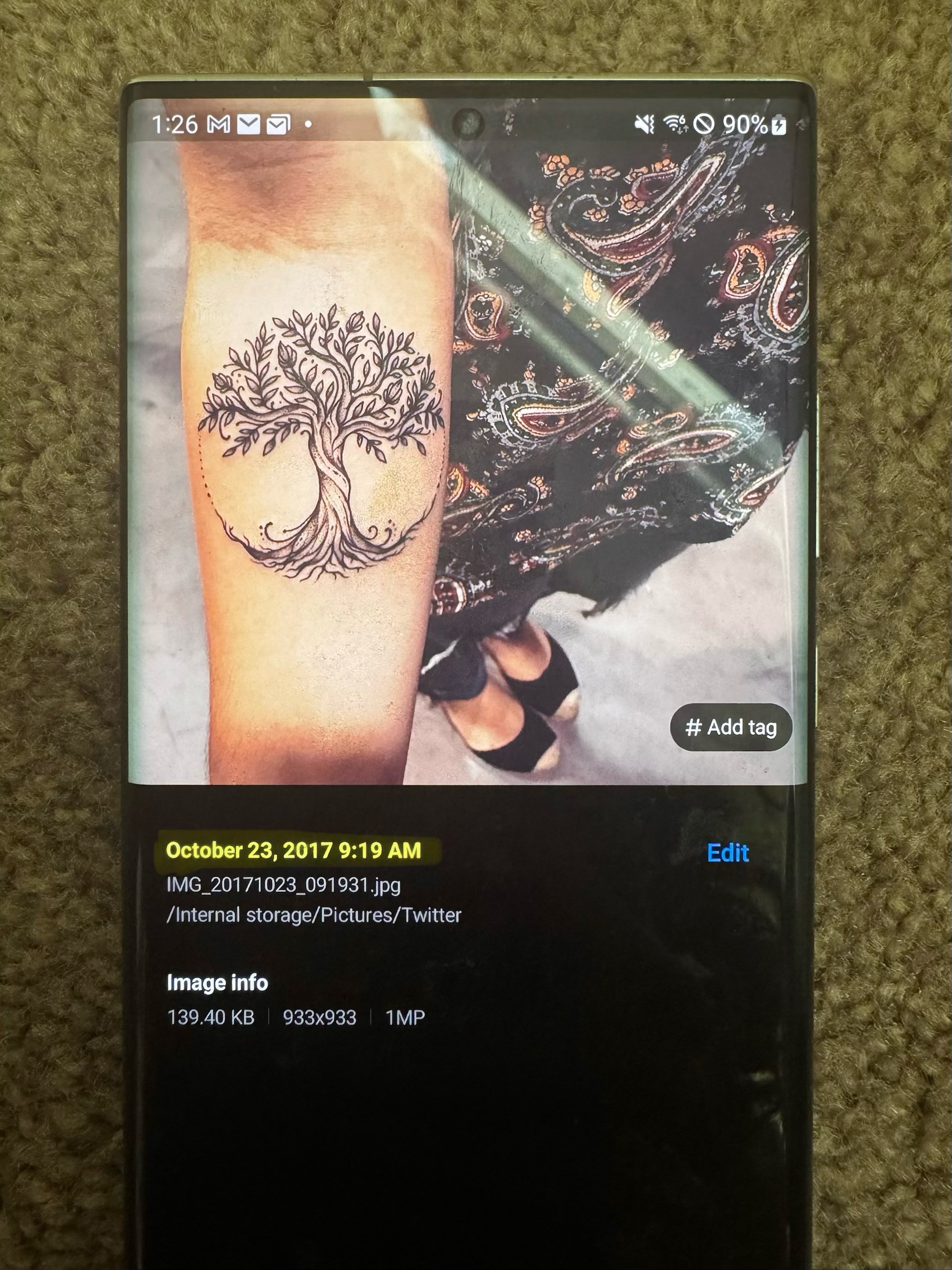 A phone screen showing a saved 2017 screenshot of a Tree of Life tattoo on someone’s forearm, with the file details dated October 23, 2017.
