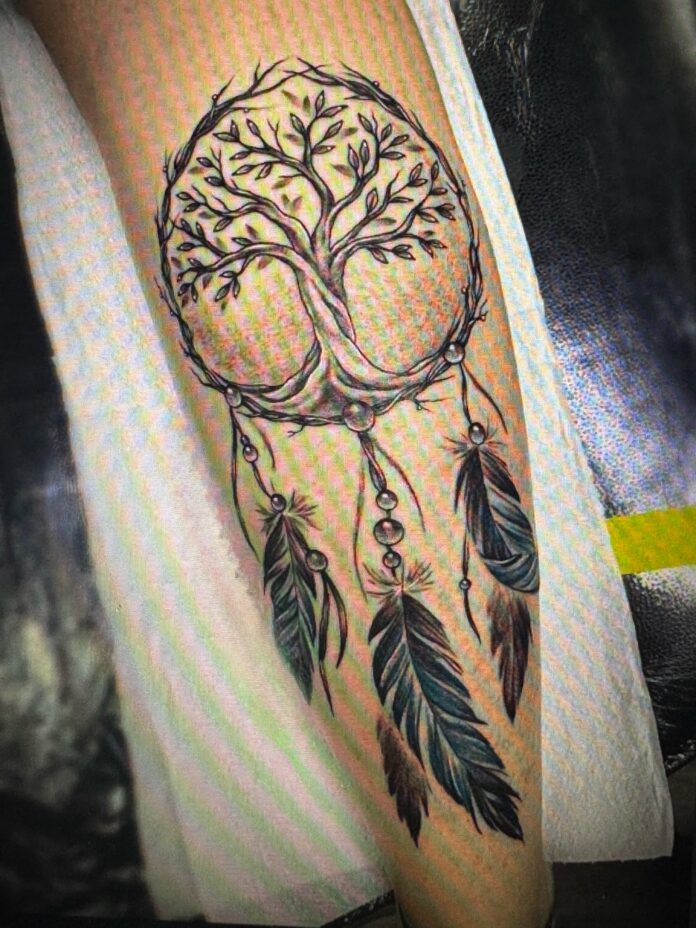 A freshly inked Tree of Life tattoo set in a circle, with feathers and beads hanging below it like a dreamcatcher, from an essay about how the author manifested a tattoo.
