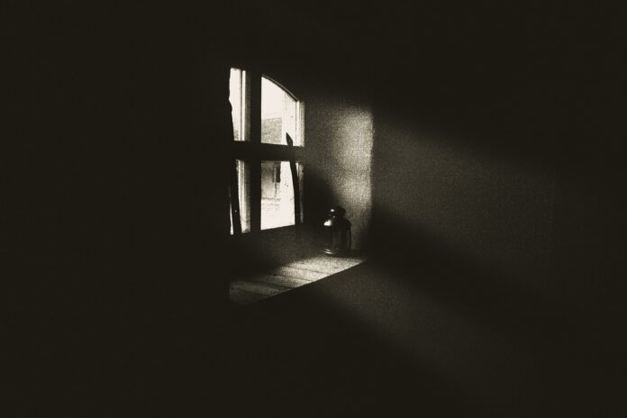A dark room where a single shaft of light from a paned window falls across the floor and wall, illustrating an essay about being Black and depressed.